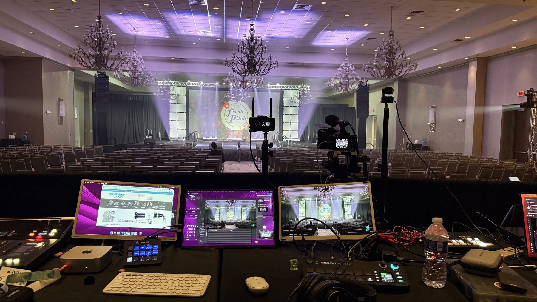 Front-of-house production desk in a hotel ballroom: three monitors showing preview, program and control, two Stream Decks, an audio interface, and two cameras on tripods facing a lit stage.