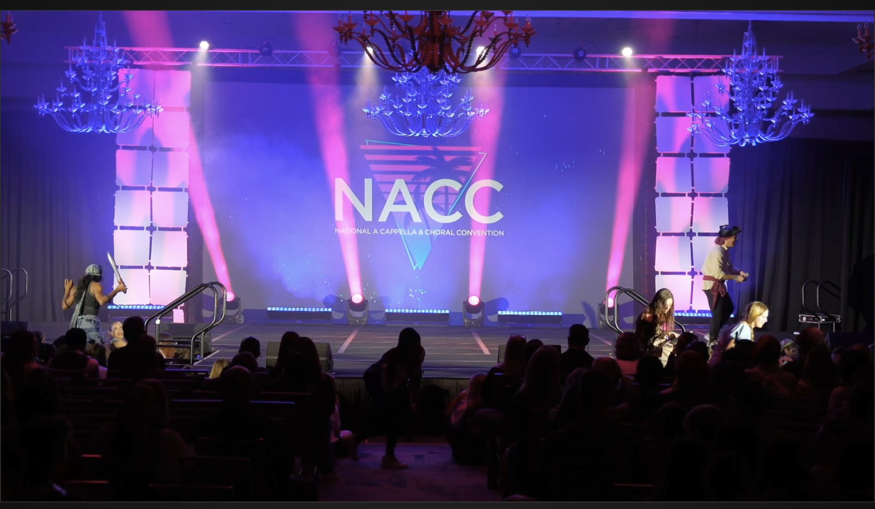 Wide shot of the full house from the broadcast: stage lit in pink and blue, the NACC screen behind, audience filling the room.