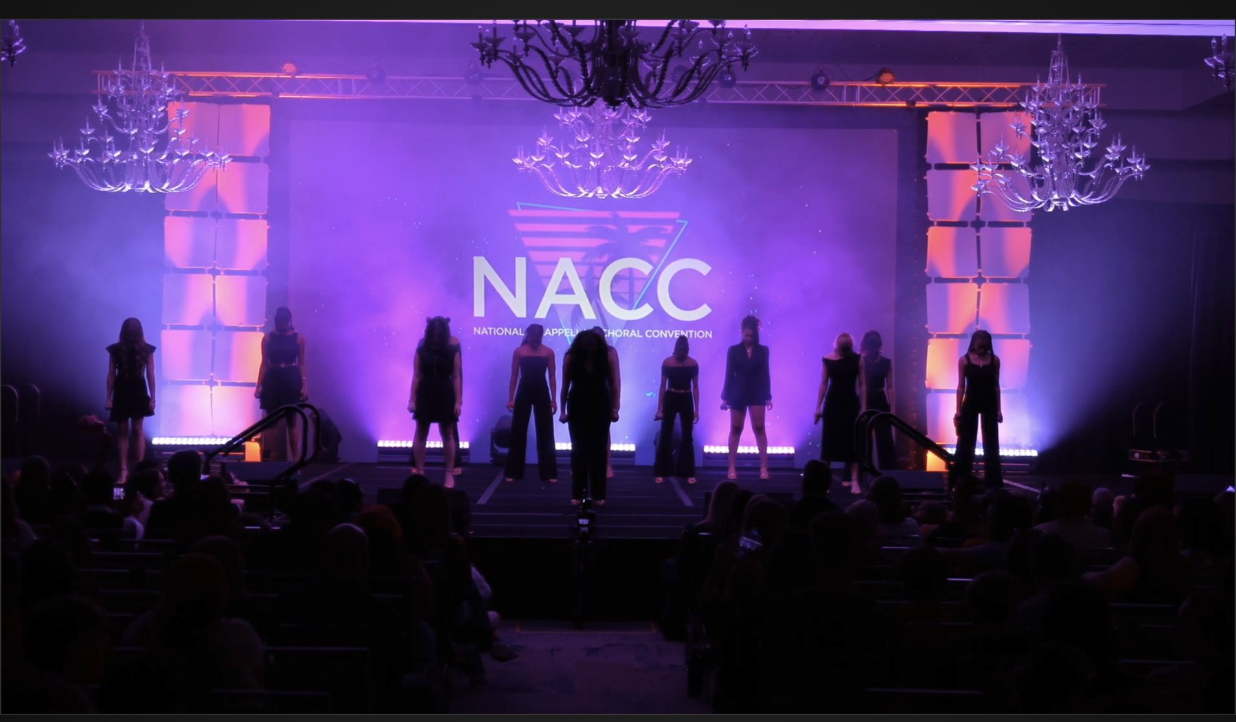 The live broadcast: a choir standing in silhouette across a stage under purple and magenta wash, the NACC logo on the screen behind them, a full audience in front.