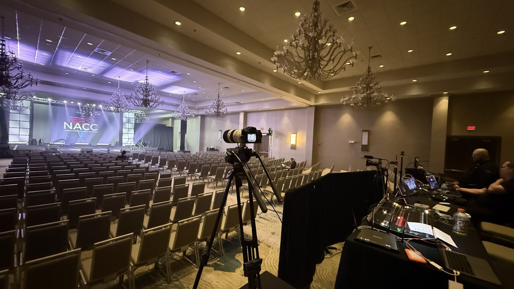 Empty ballroom set for a convention, rows of chairs facing a stage with the NACC logo on a large screen, a camera on a tripod mid-room.