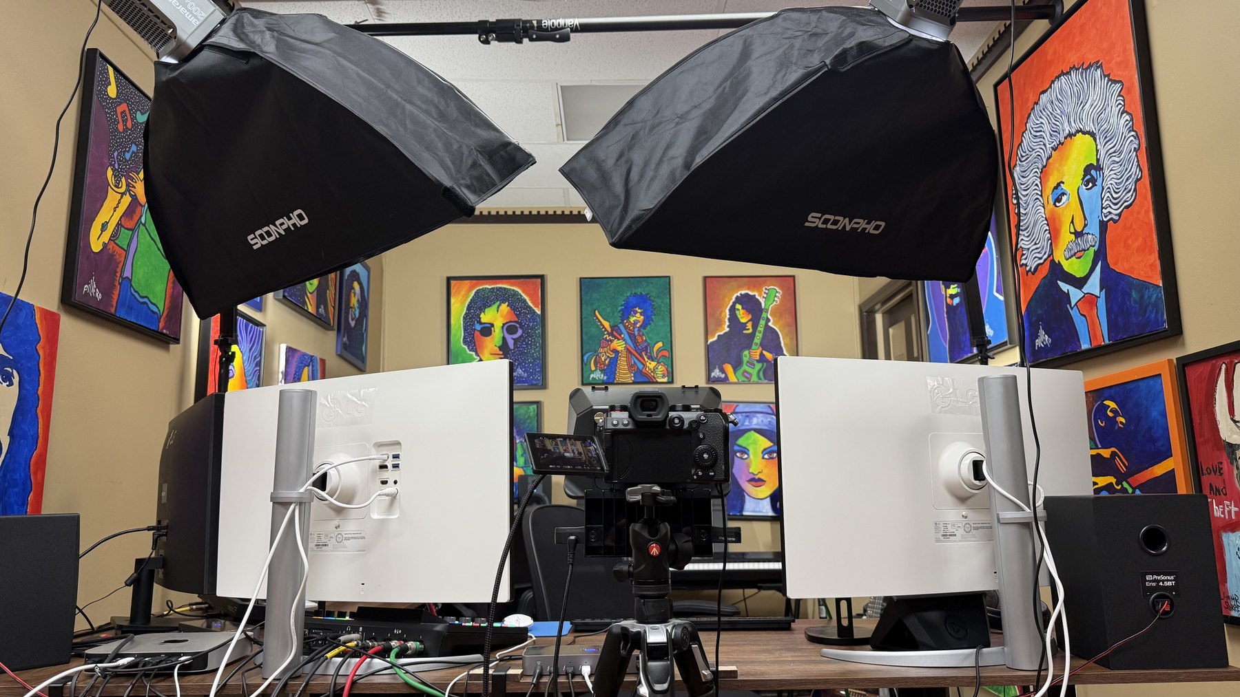 Seen from behind the desk: a single mirrorless camera on a tripod, centred at eye height between two monitors, its flip-out screen showing the shot.