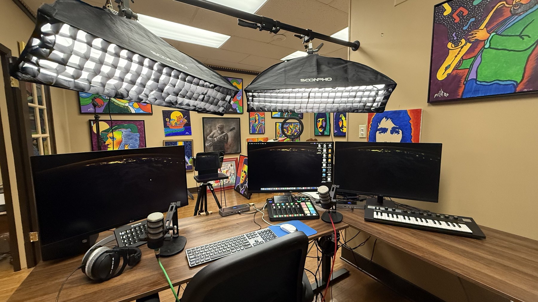 The same studio, now clear: two curved monitors either side of a centre screen, a compact mixer, keyboard and microphone on an uncluttered desk under two softboxes.