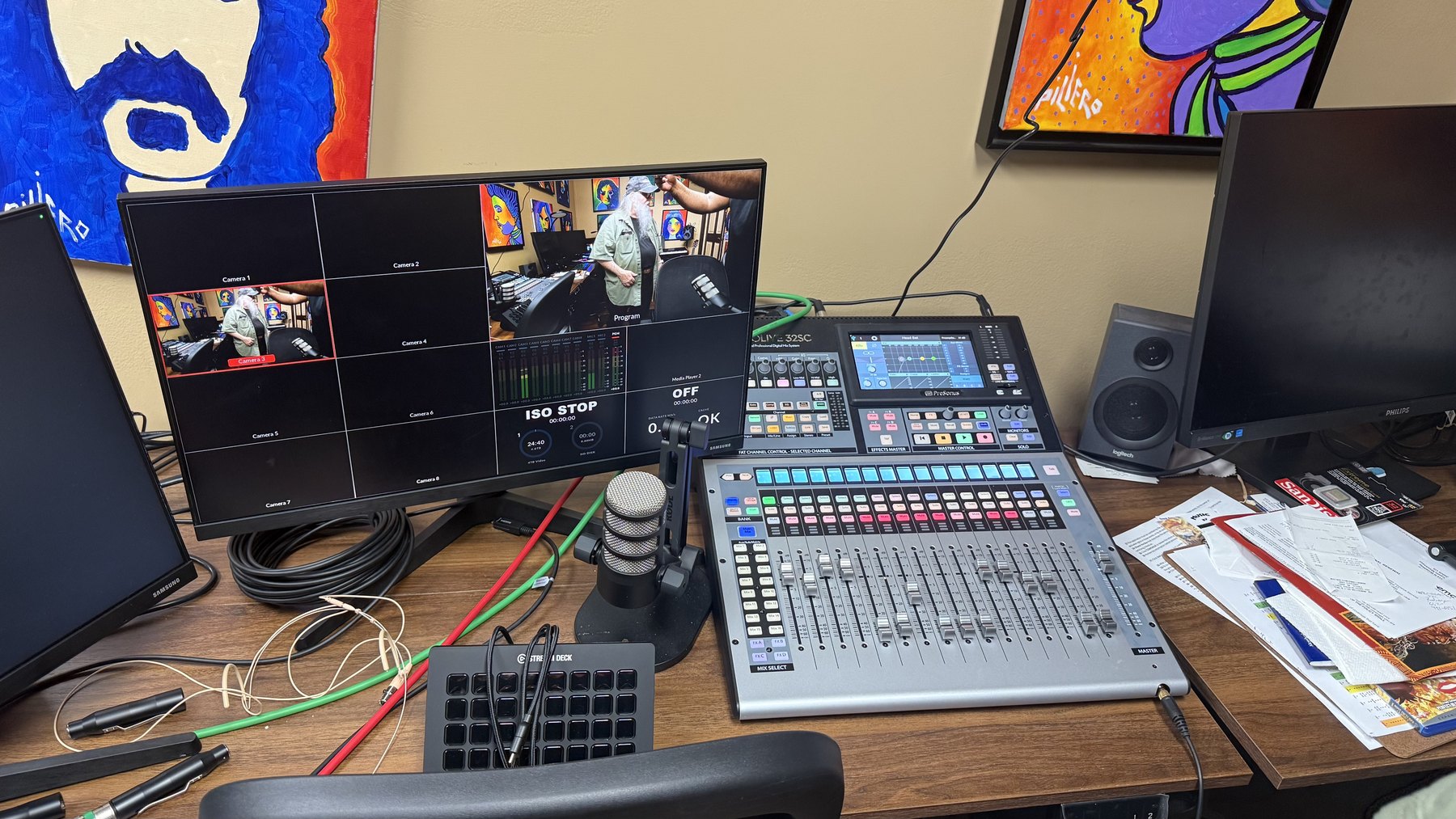 A production desk: a monitor showing a grid of camera slots beside a large 32-channel digital mixing console, a Stream Deck, coiled cable and stacked paperwork.