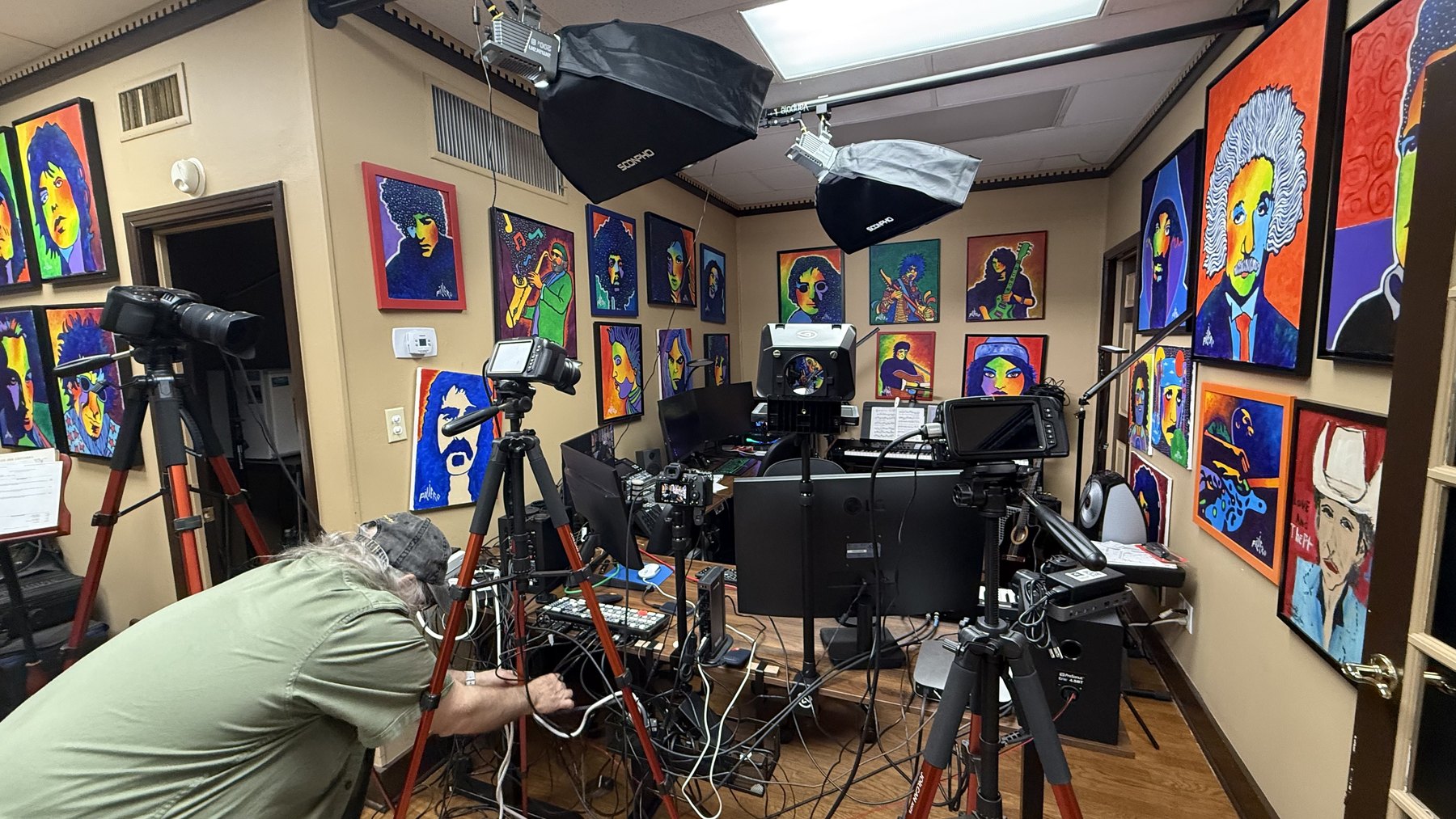 A small studio crowded with camera tripods, softboxes and cable running across the floor, walls covered in brightly coloured painted portraits.